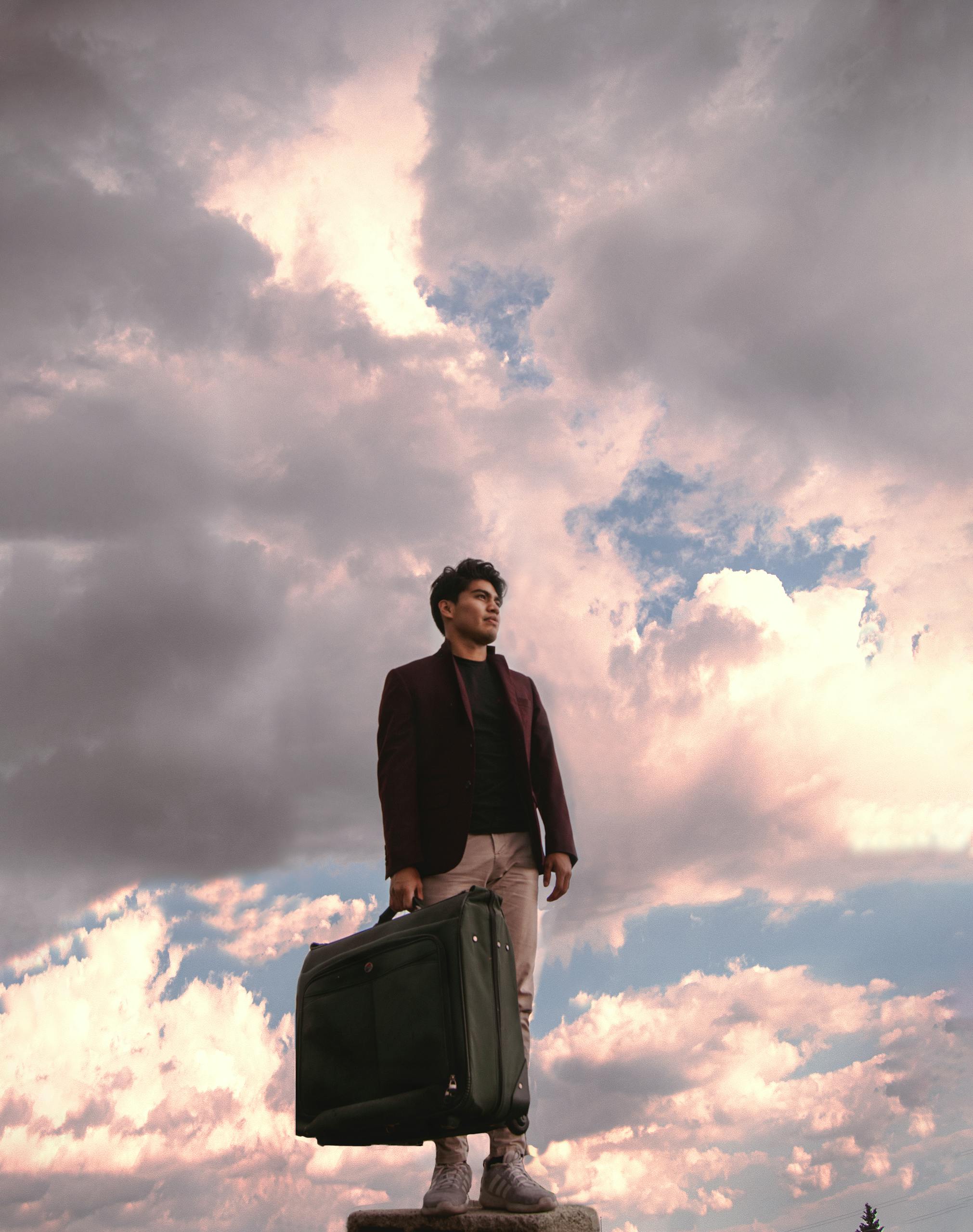 A man stands confidently with a suitcase against a dramatic, cloudy sky.
